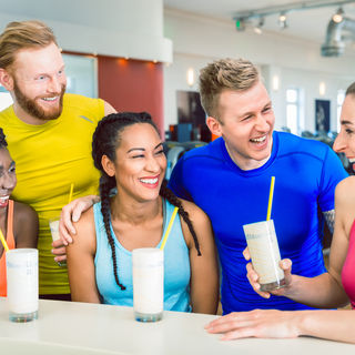 Das Bild zeigt eine Gruppe von 5 Sportlern in einem Fitness Studio, drei Frauen und zwei M&auml;nner, jeweils in einem sportlichem Outfit, welche einen Eiwei&szlig;shake an der Theke/Servicebereich in einem Fitness Studio genie&szlig;en.